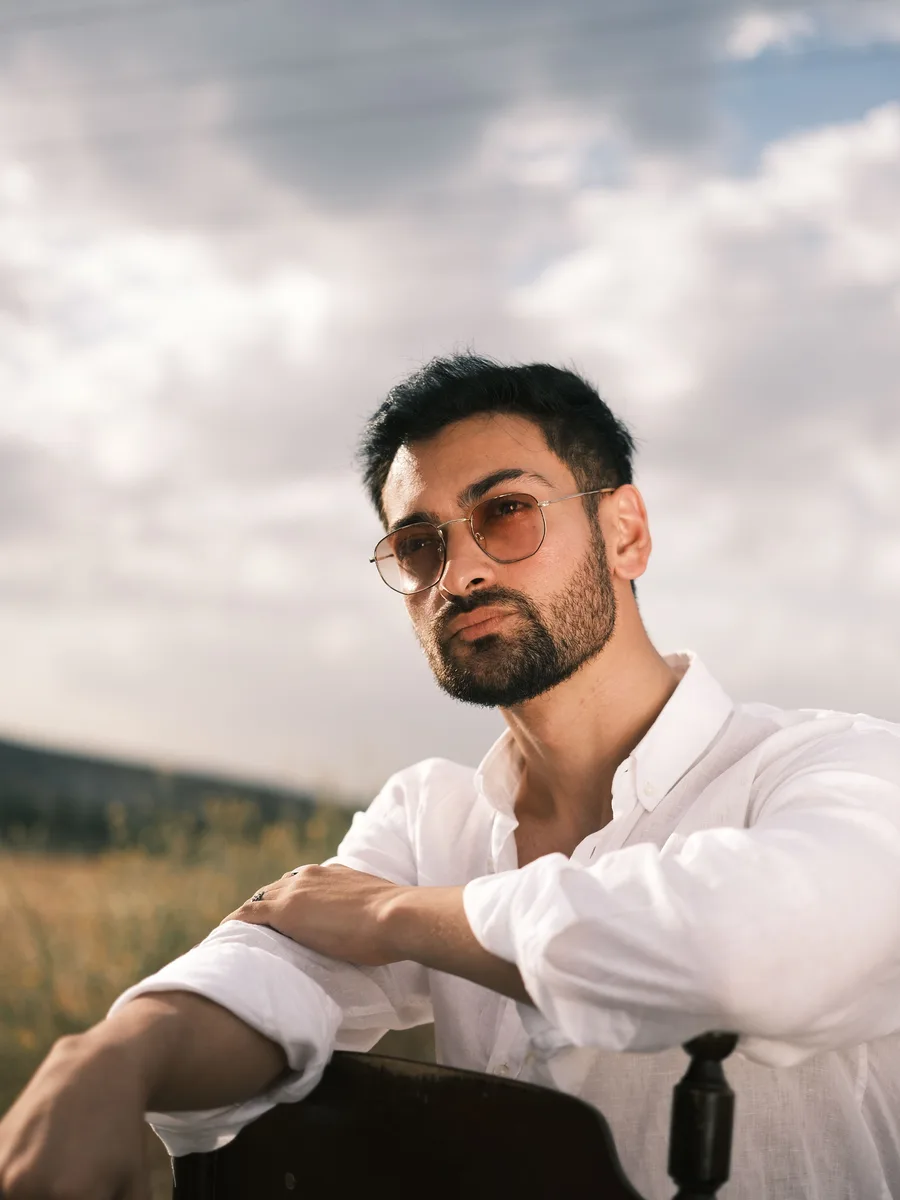 Original photo — a man in a white shirt and round sunglasses seated outdoors under a cloudy sky